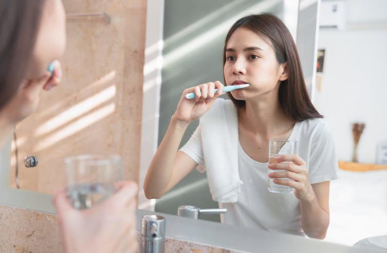 Young woman brushing teeth in bathroom mirror, holding glass of water