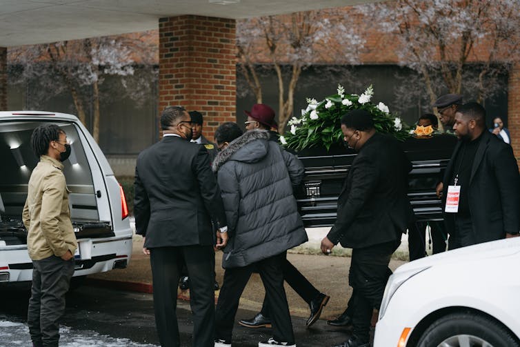 Mean wearing black and gray carry a black casket topped with white flowers to the open back of a white hearse.