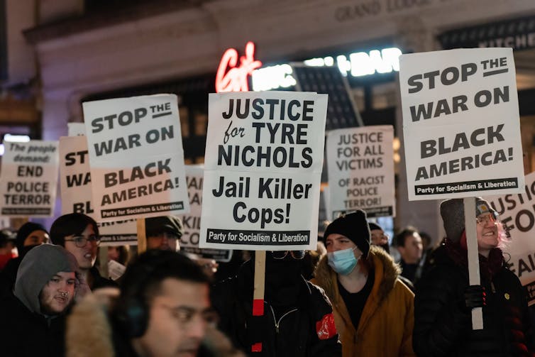 People wearing winter coats and hats protest outside holding black and white signs.