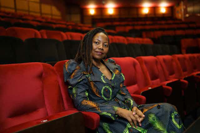 A woman in a dress of African fabric sits in a theatre with red chairs, looking directly to camera with a half smile.