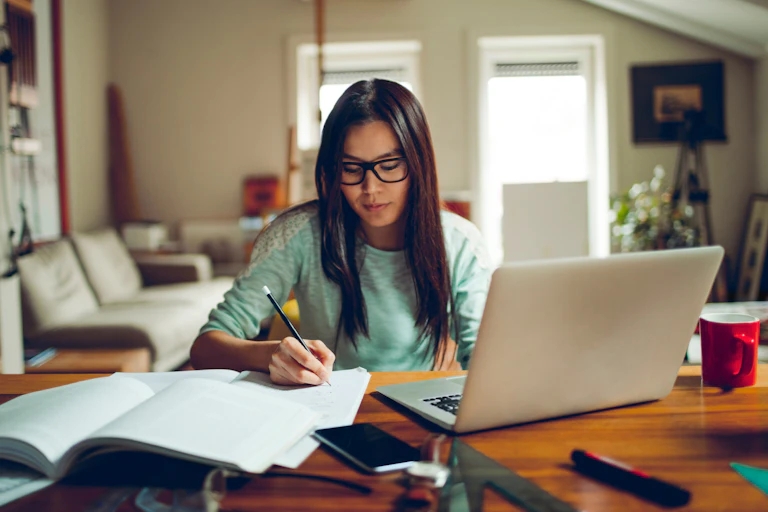 Girl takes notes as she sits in front of a laptop at a desk.