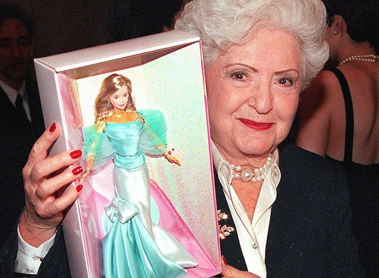 Elderly woman with white hair wearing necklace and red lipstick holds box containing a doll wearing a turquoise and pink dress.