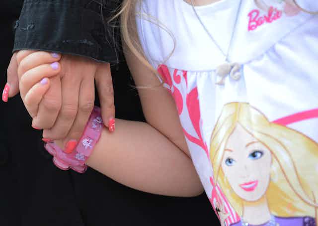 Close-up shot of woman holding hand of daughter, who's wearing a pink watch and a shirt with Barbie's likeness on it.