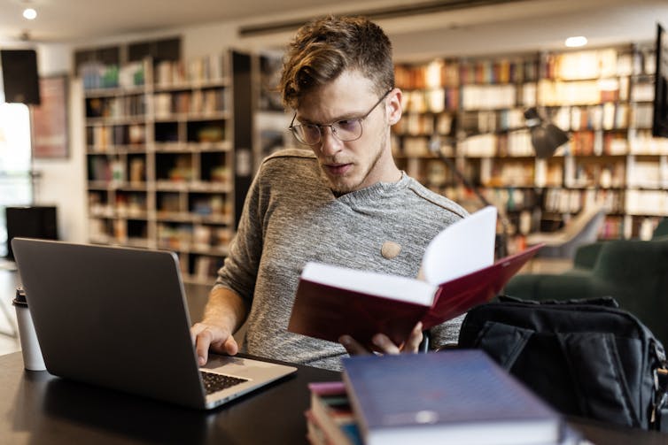 A man with textbooks and a laptop works in a library.