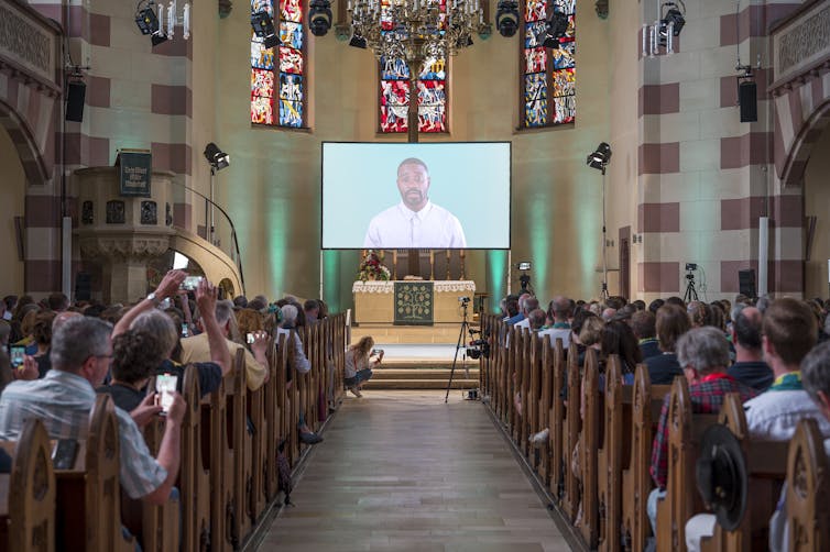 People sitting on either side of the pews while an avatar on a screen in front delivers a sermon.