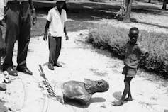 Black boys stand beside a toppled statue in a black and white photo from the 1960s.