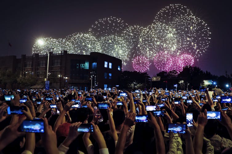 Thousands of people at a night-time celebration hold up their smartphones to take pictures of fireworks.