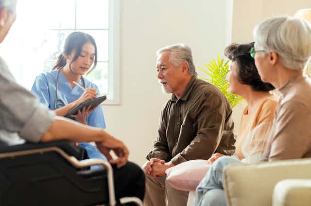 A group of older people sit in a circle and talk with a medical professional who has a clip board.