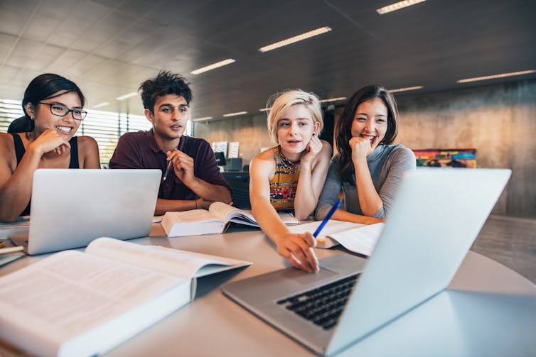 Four students work at a desk with books and laptops.