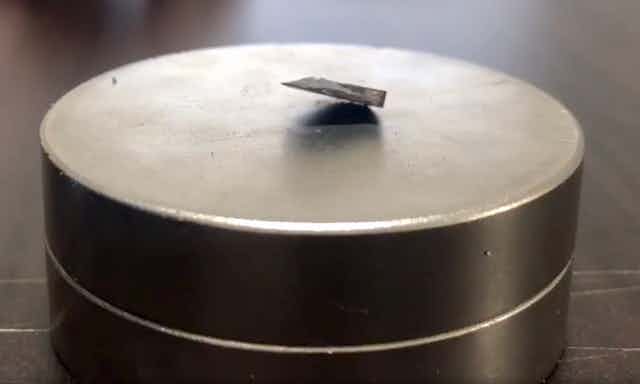 A photo showing a chip of dark material partially floating above a silver metallic magnet.