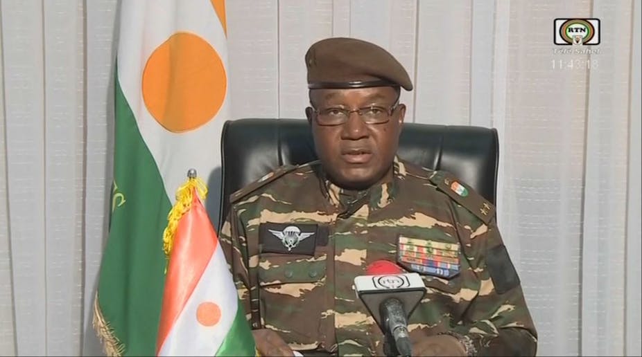 A man in military fatigues speaking into a microphone while seated on a black chair, with two flags with orange, white and green stripes and an orange dot in the middle behind him and to his right