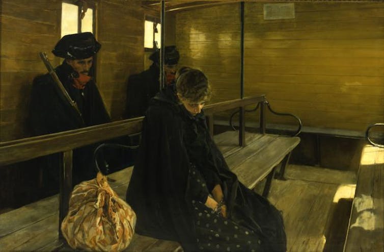 A woman sitting on a wooden bench in a train carriage, dressed in black, gazes at the ground under the watchful eye of a civil guard.