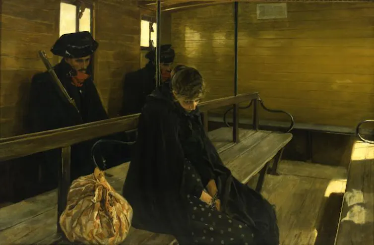 Una mujer sentada en un banco de madera de un vagón de tren y vestida de negro, mira al suelo ante la vigilancia de un guardia civil.