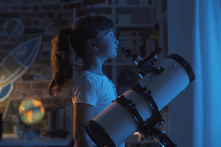 Girl standing by telescope looking out of window at night