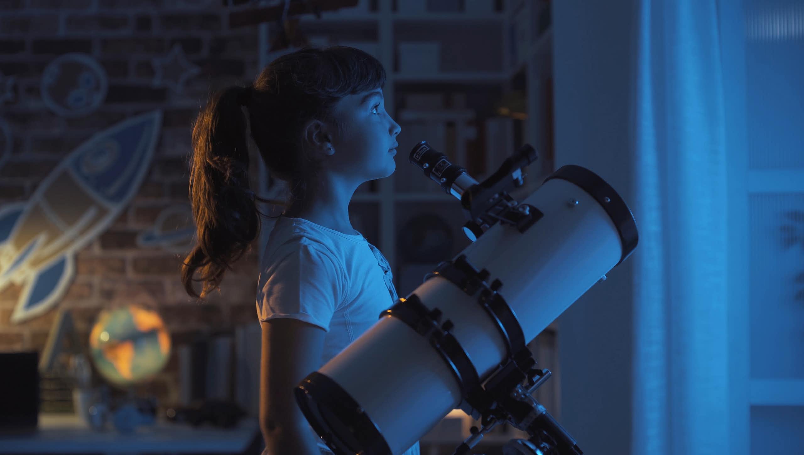 Girl standing by telescope looking out of window at night