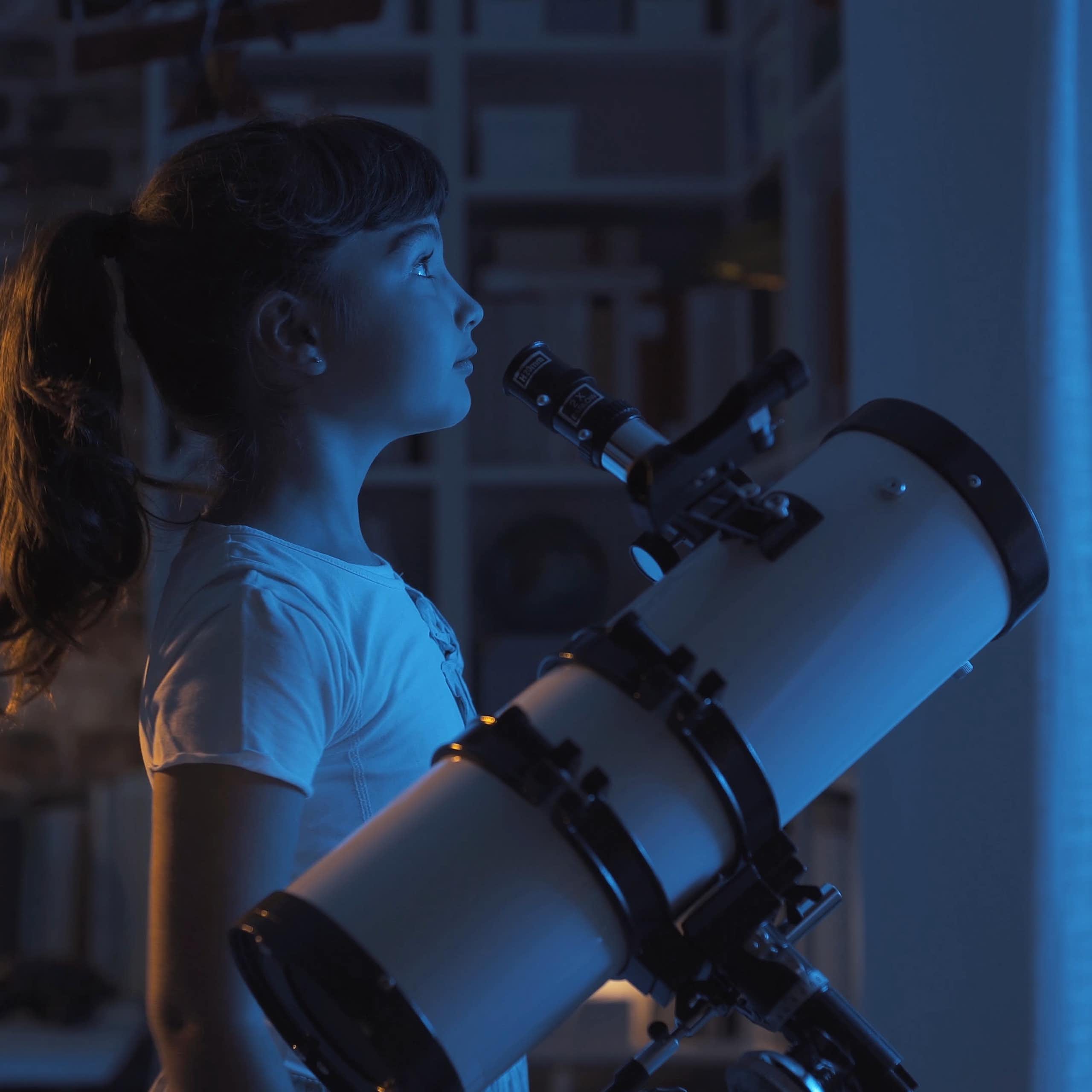 Girl standing by telescope looking out of window at night