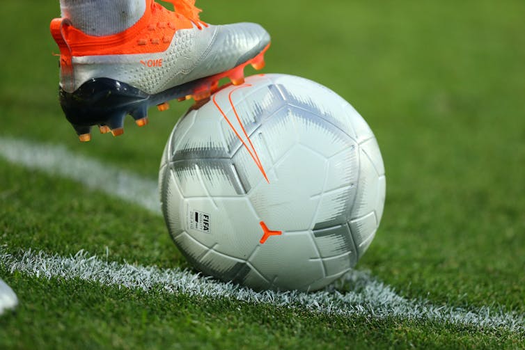 A foot in a football boot resting on a football on a grass pitch.