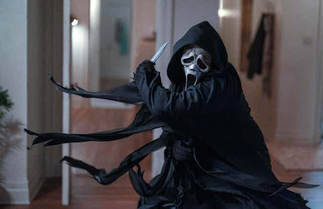 Figure in black hooded robe and scream mask slashing with a knife in an apartment.