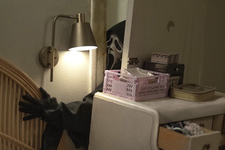 A knife-weilding figure in skinny skeleton mask busts through a bedroom door with a messy dresser jammed up against it.