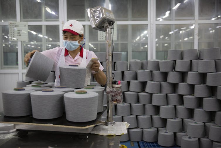 A person wearing a face mask and work uniform picks a large spook of yarn up from a pile