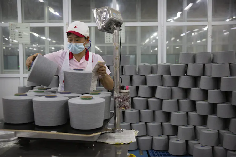 A person wearing a face mask and work uniform picks a large spook of yarn up from a pile
