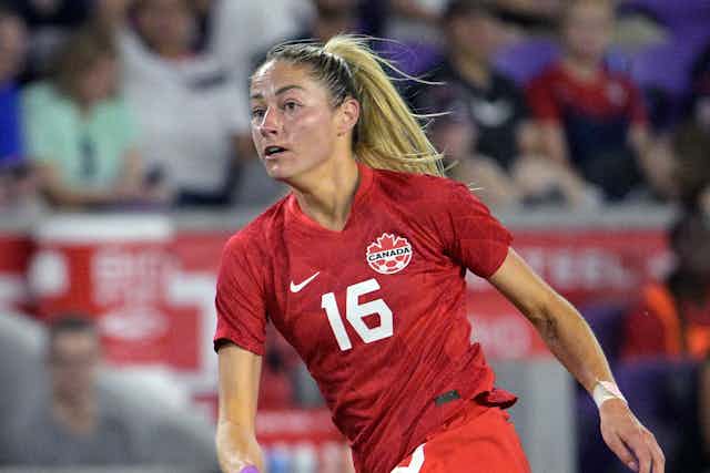A picture of a blonde woman from the waist up, wearing a Team Canada soccer jersey and looking down a soccer field
