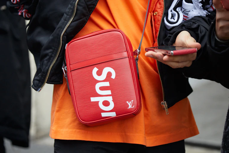 Woman wearing red Louis Vuitton crossover bag showing Supreme streetwear label.