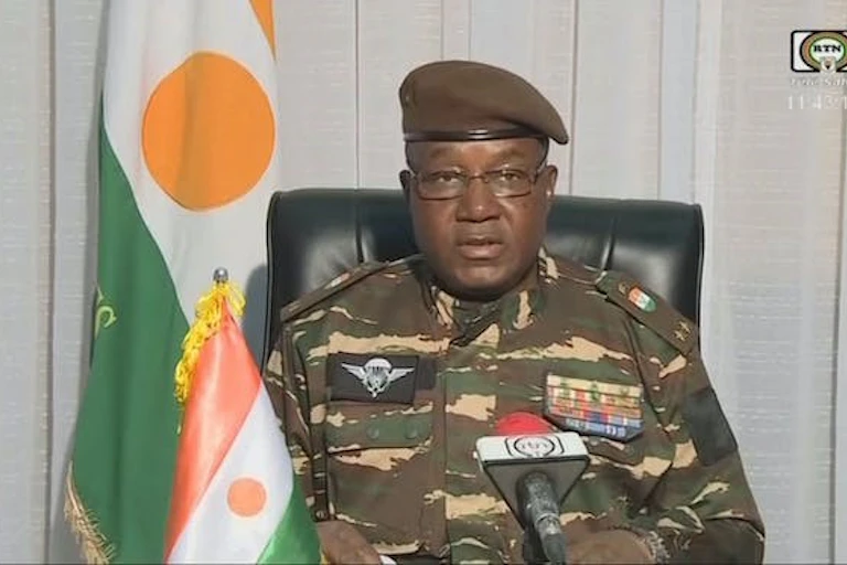 Un homme en treillis militaire parle dans un microphone, assis sur une chaise noire, avec deux drapeaux à bandes orange, blanches et vertes et un point orange au milieu derrière lui et à sa droite.