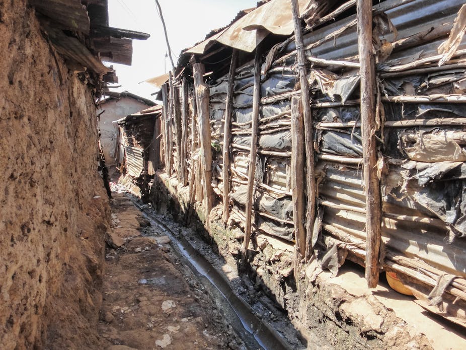 A stream of water running through a makeshift drain between two structures - one built with slim strips of wood and plastic bags, and the other with mud; both have iron sheet roofs