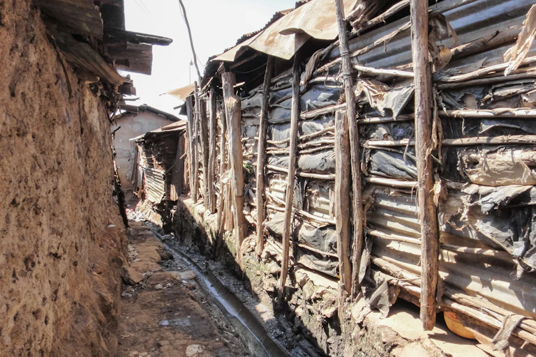 A stream of water running through a makeshift drain between two structures - one built with slim strips of wood and plastic bags, and the other with mud; both have iron sheet roofs