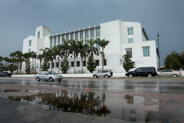 A large white building is seen with palm trees and streets covered with puddles.