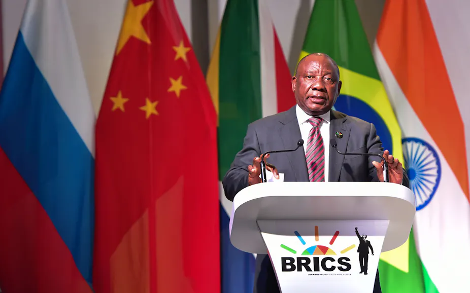 A man wearing a suit and tie speaks at a podium bearing the BRICS logo.