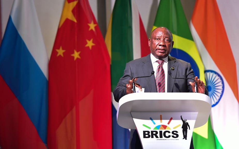 A man wearing a suit and tie speaks at a podium bearing the BRICS logo.