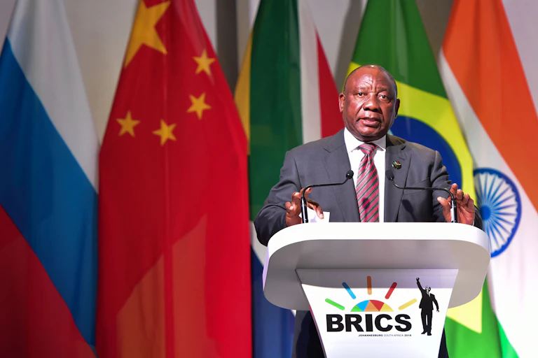 A man wearing a suit and tie speaks at a podium bearing the BRICS logo.