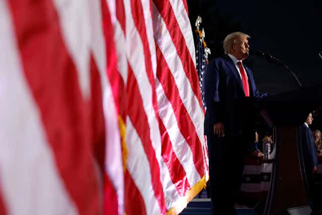 Donald Trump stands on a stage in a dark suit in front of a row of American flags.