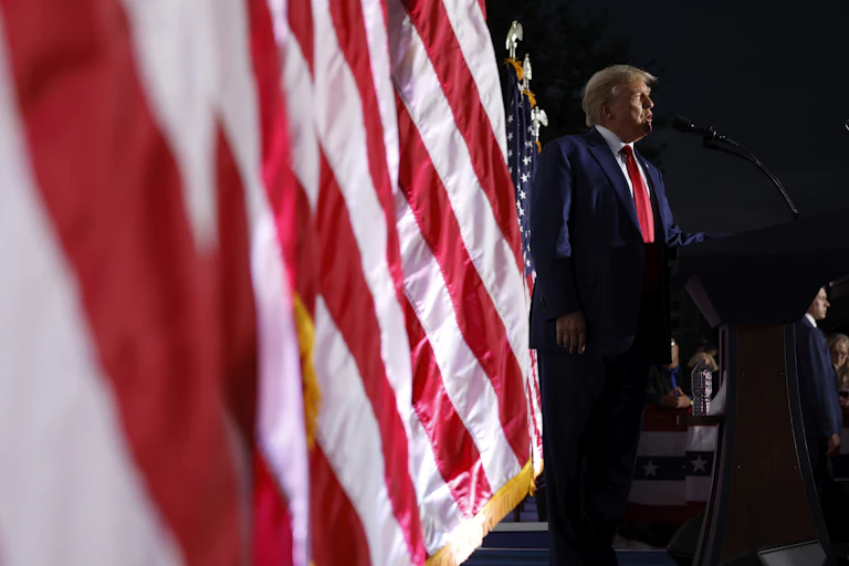 Donald Trump stands on a stage in a dark suit in front of a row of American flags.