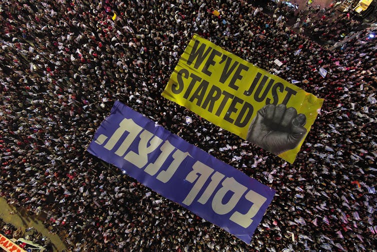 A large crowd holding a sign in Hebrew and English that shows a fist and says 'We've just started.'