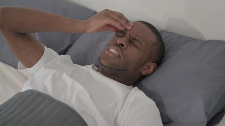 A man lying in bed with a pained expression, with his hand to his head