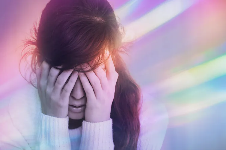 A woman with her hands covering her face, with streaks of coloured light around her head