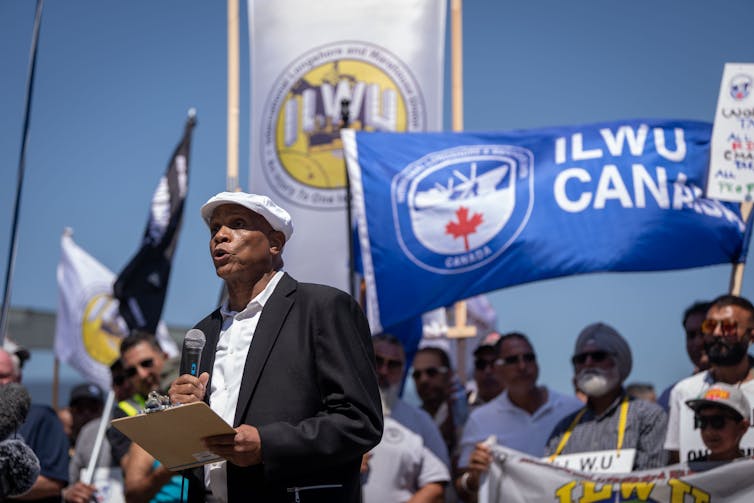 A Black man in a blazer and newsboy cap speaks to a crowd of people waving flags and holding signs that say ILWU CANADA