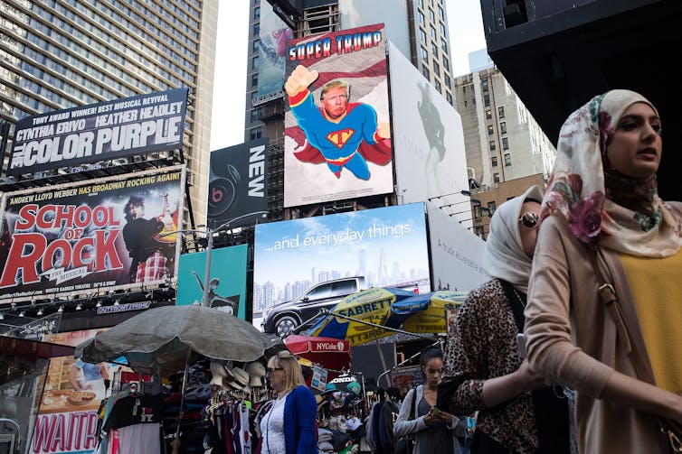 People walk through Times Square, in front of a large poster that says 'Super Trump' and shows Trump's face on a Superman body, flying.