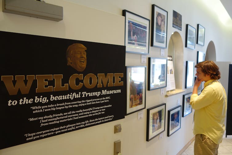 A man looks at a wall covered in photos and text, including one plaque that says 'welcome to the big, beautiful Trump museum.'