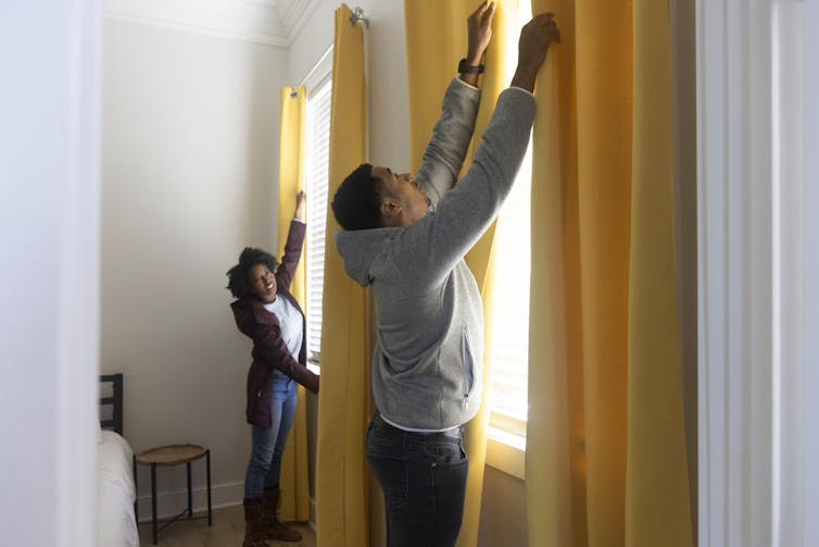 A man and woman close bright-yellow curtains on tall side-by-side windows. The windows also have shades.