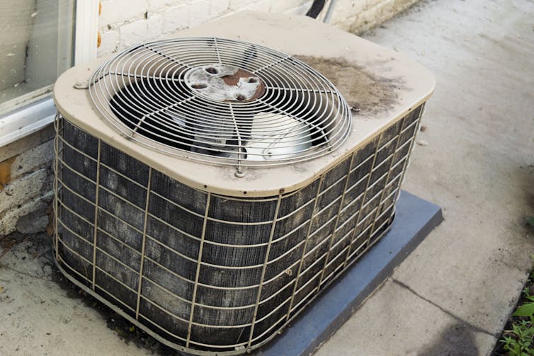 An old, dirty and beat-up air condition sits next to a window and brick wall with peeling paint.