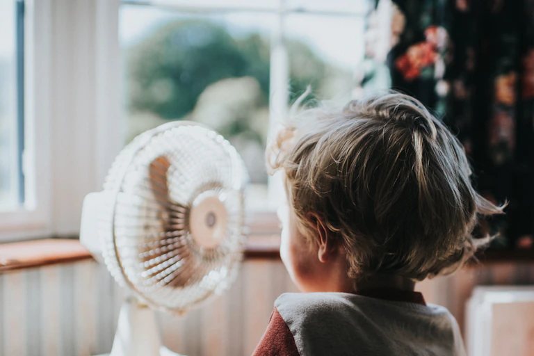 A boy stands in front of a fan with light coming in older windows behind it.