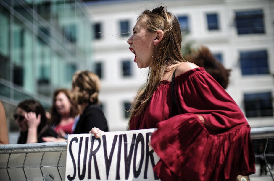 A woman whose mouth is wide open in a scream, wears a red blouse and holds a sign that reads, “Survivor.”