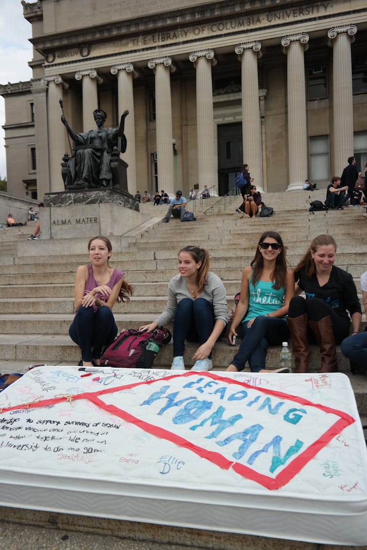 Four casually dressed women sit on a building’s outside stairs. A mattress with messages scrawled on it rests on the stairs before them.