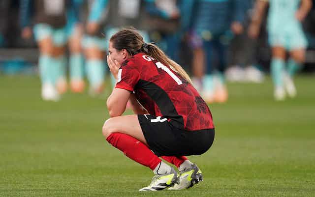 A woman in a red and black soccer uniform crouches on a soccer field with her hands over her mouth