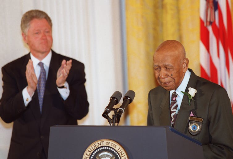 A Black man is speaking into a microphone near the American flag as a white man dressed business suit applauds.