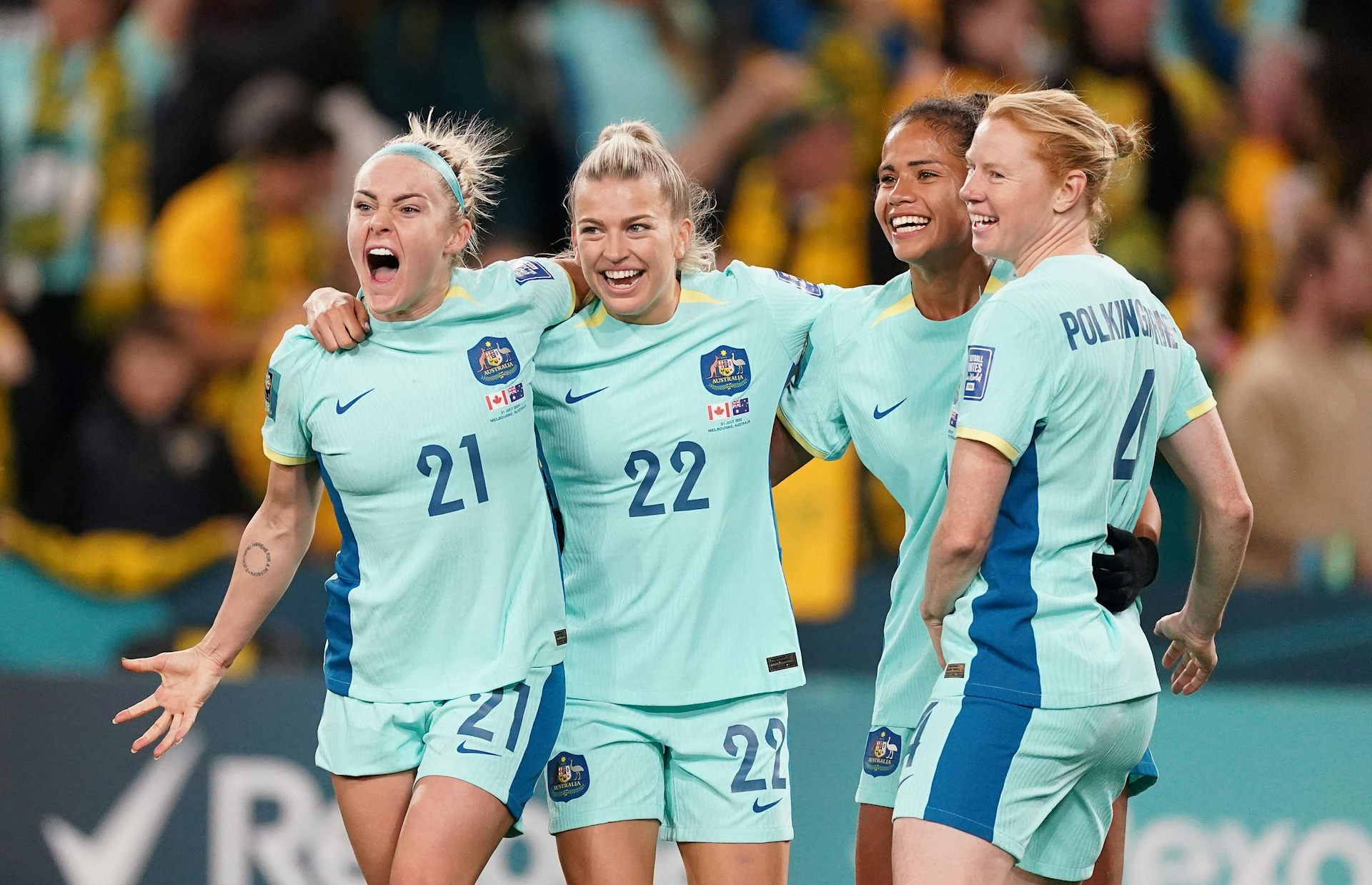 Four women in pale turquoise soccer uniforms stand with their arms around one another, looking excited